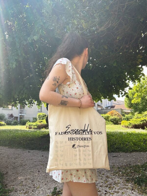 Tote bag Pluma Flora porté à l’épaule par une jeune femme dans un jardin ensoleillé, avec le message “Ensemble faisons briller vos histoires”