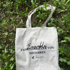 Tote bag en coton naturel Pluma Flora sur fond de verdure, avec message "Ensemble faisons briller vos histoires"