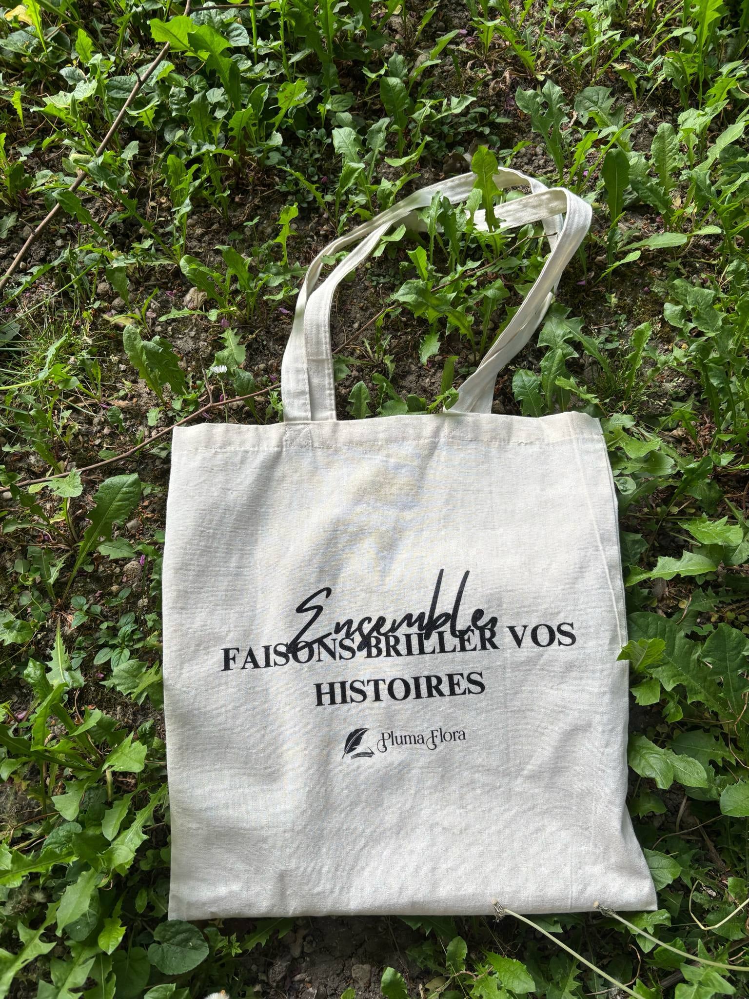 Tote bag en coton naturel Pluma Flora sur fond de verdure, avec message "Ensemble faisons briller vos histoires"